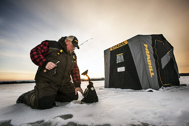 A fisherman squats over an ice fishing hole, holding an ice rod in his hand.