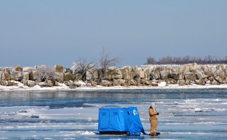 A fisherman stands ice jigging on a frozen lake beside his snow shelter.